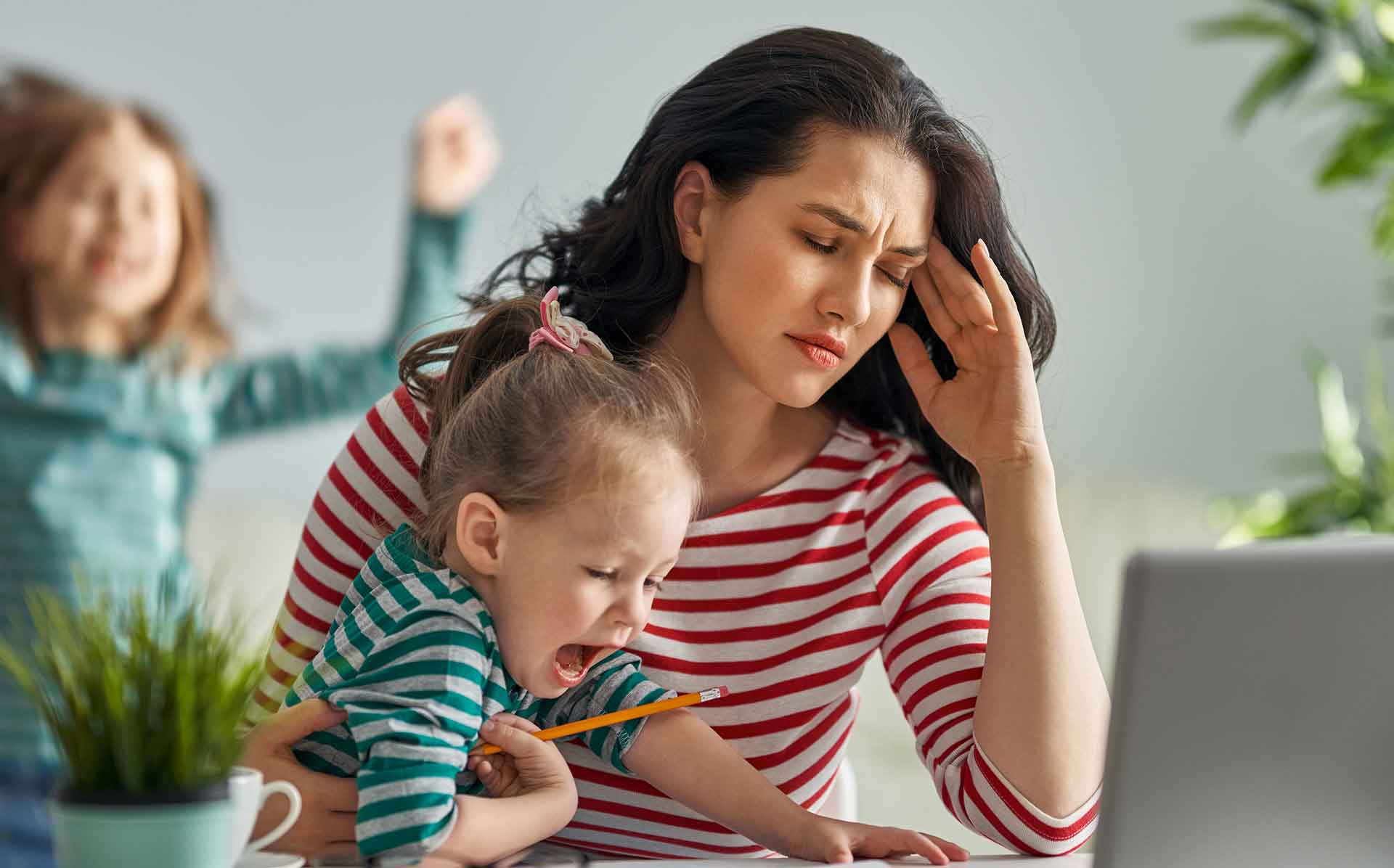 A woman with a headache holding a baby with a child behind her